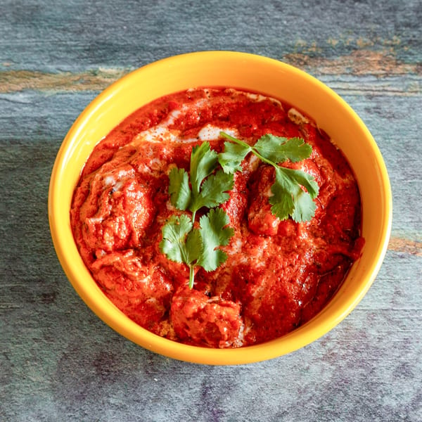 Vibrant red butter chicken in a yellow bowl, garnished with fresh cilantro, on a rustic gray surface.