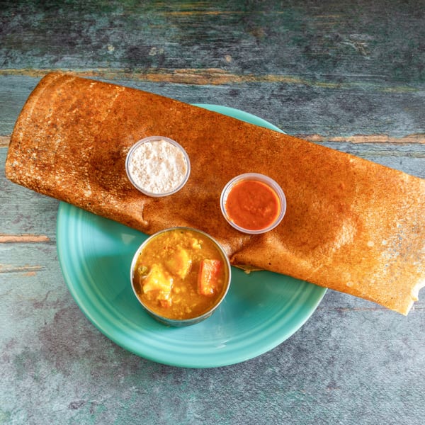 Crispy brown dosa on a teal plate with three small bowls of chutneys and sambar.